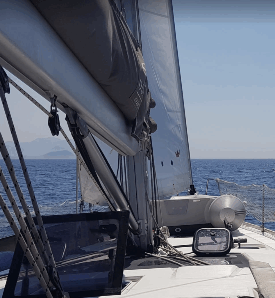 From the upper deck of a sailing boat, a picture of the deck, the sails and the horizon. On the left there is a land.